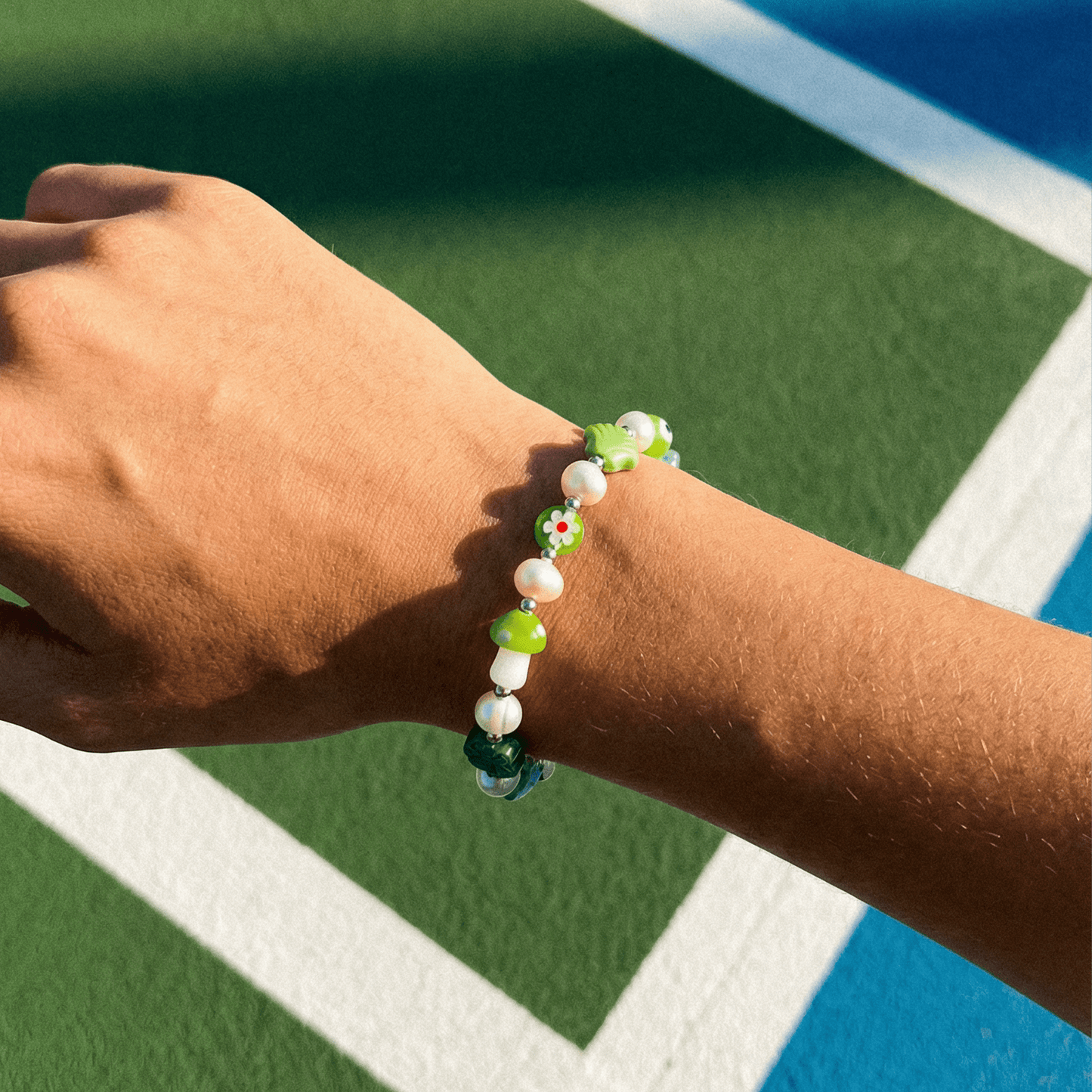 Grünes Mood Armband mit Keramik- und Perlen-Elementen am Handgelenk, getragen auf einem sonnigen Tennisplatz mit grün-blauem Boden im Hintergrund