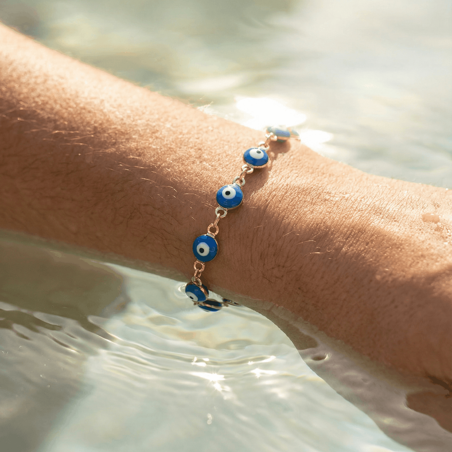 Nahaufnahme eines Handgelenks, das teilweise im klaren Wasser liegt. Am Handgelenk wird ein blaues Armband mit kleinen, runden Elementen getragen. Wassertropfen auf der Haut reflektieren das Sonnenlicht, der Hintergrund ist weich und unscharf