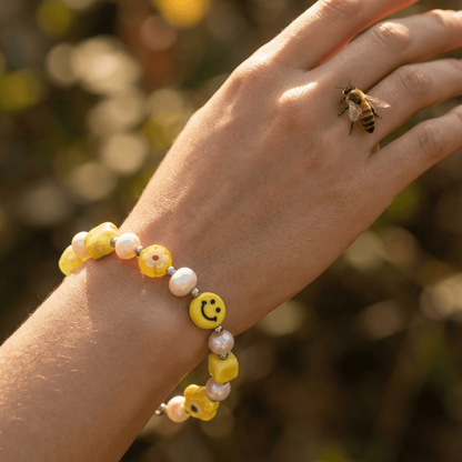 Nahaufnahme eines Frauenhandgelenks im warmen Sonnenlicht. Am Handgelenk trägt die Frau ein gelbes Armband mit Keramikperlen, Süßwasserperlen und einem Smiley-Element. Auf einem Finger sitzt eine kleine Biene. Der Hintergrund ist unscharf und zeigt natürliche, grün-goldene Farbtöne