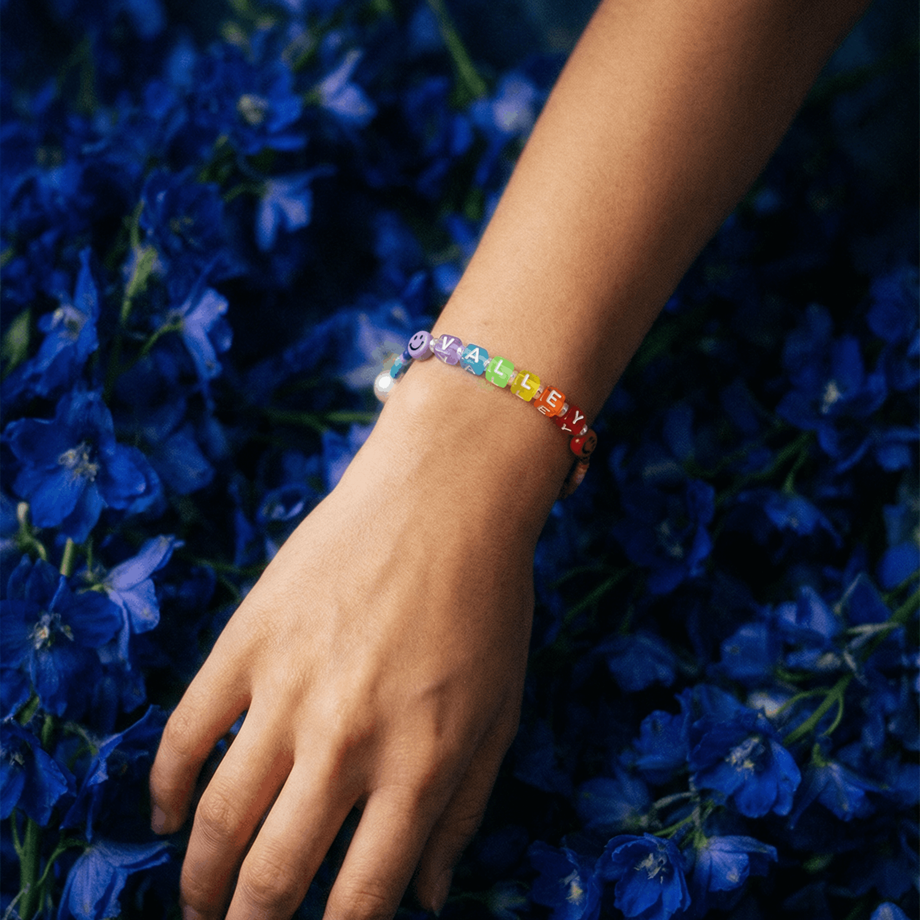 Personalisiertes Armband mit bunten Buchstaben-Perlen am Handgelenk, getragen über einem dichten Hintergrund aus tiefblauen Blumen im natürlichen Licht