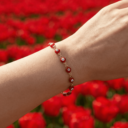 Rotes Spirit Armband mit kleinen Augen-Perlen am Handgelenk, getragen vor einem unscharfen Hintergrund aus leuchtend roten Blumen im natürlichen Licht
